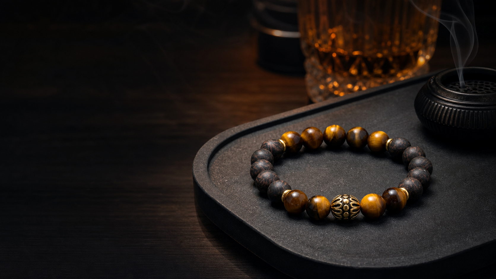CENSEROOT Black Accord wearable solid incense bracelet on a dark desk, with tiger eye beads, black incense beads, and warm amber light.
